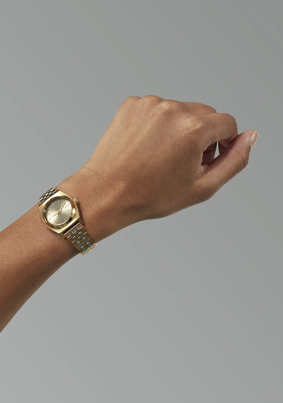 A gold-tone watch with a matching metal link bracelet worn on a person's wrist, shown against a plain gray studio background.